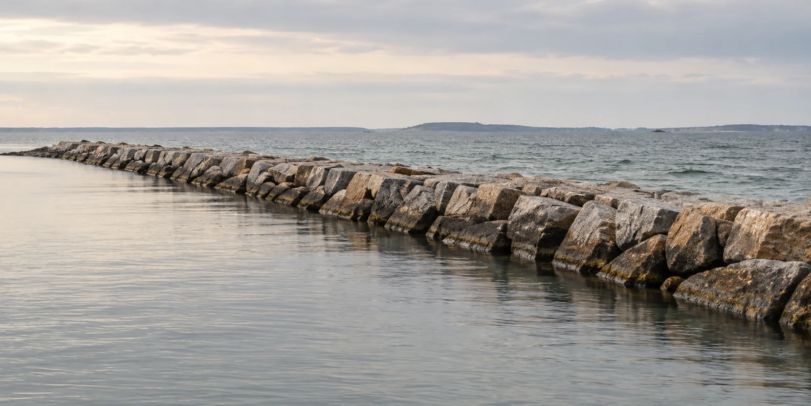 A granite breakwater curving out into calm water under a pale sky, a low headland on the far horizon