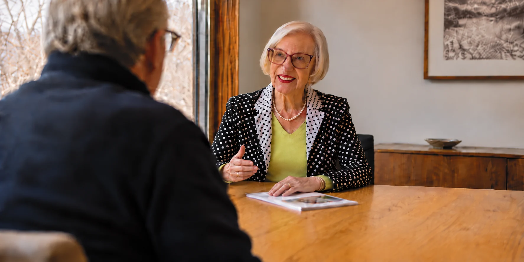 Two people in conversation across a light oak table beside a large window, one speaking and gesturing as the other listens